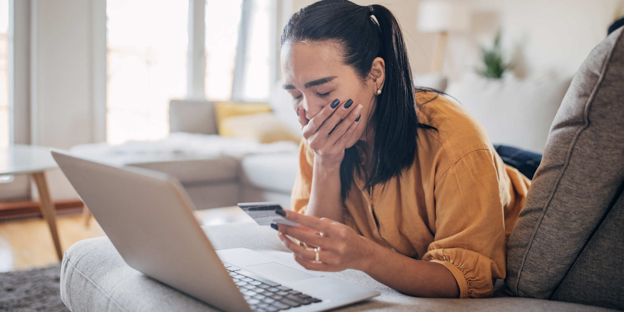 Dark-haired woman with hand over mouth looking down at credit card and laptop screen