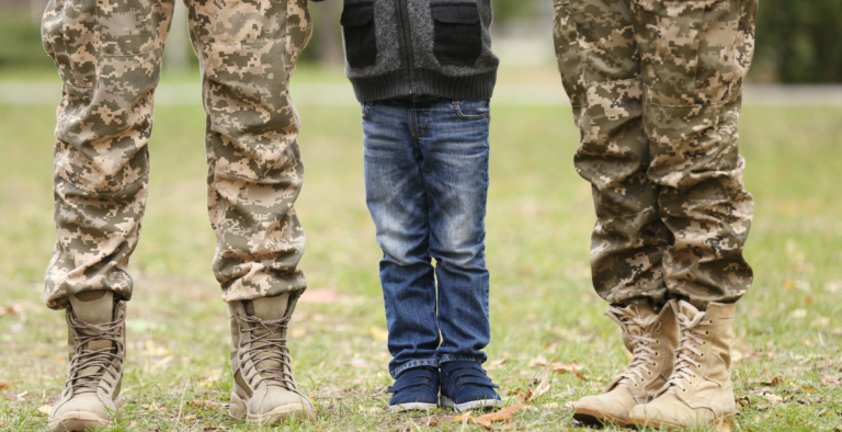 A military family standing together outdoors, depicting the legs of two armed forces members in uniform alongside a child.